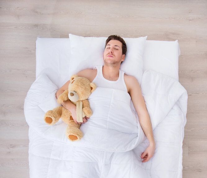 Dude learns life lessons as he sleeps on the dining room floor, wearing a white tank top and holding a teddy bear. 